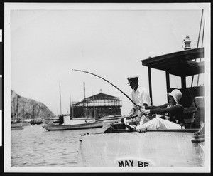 A woman fishing from a boat, with a man at her side, ca.1920