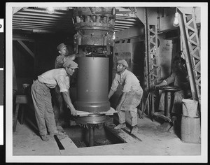 Unidentified factory interior, showing workers adjusting a small slab beneath a large machine, ca.1930