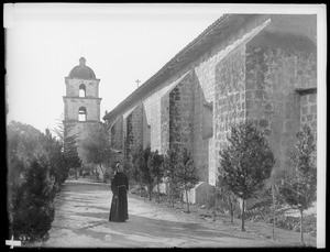 Monk on a walkway that leads into the cemetery at the Santa Barbara Mission, 1898