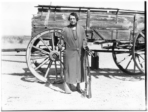 Old woman holding rifles and revolvers in front of a wagon