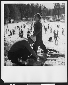 Man helping a boy put on skis at Big Pines mountain camp, ca.1940