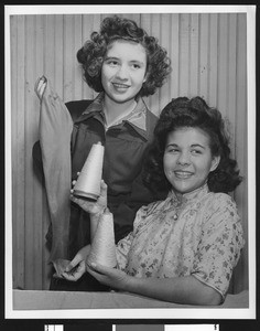 Portrait of two female garment workers holding two spools of thread and fabric in a pose for the camera, ca.1940