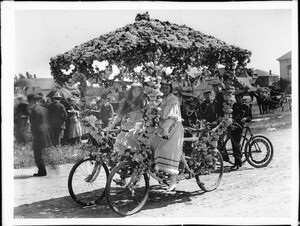 Two women sitting on a flower festooned double tandem (side by side) cart in the Los Angeles Fiesta, ca.1901-1905