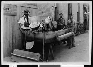 Exterior view of a sewer maintenance yard, showing three workers in protective masks standing next to the "sewer skiff", ca.1936