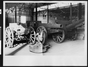 Three artillery pieces and a mortar displayed inside a large warehouse, ca.1917