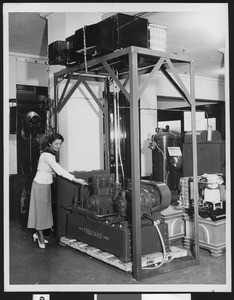 Unidentified factory interior with a woman posing next to a "Super Frigidaire Power..." piece, ca.1950