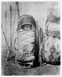 Portrait of an Apache papoose tightly wrapped in a papoose basket, strapped to a carryall, and propped up next to a bow and arrows, Arizona, 1880