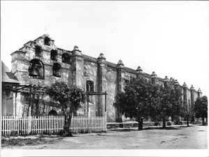 View of the front of Mission San Gabriel lined with pepper trees, ca.1886