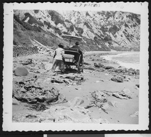 Woman walking behind a carriage along the rocky shore of a beach between Ventura and Santa Barbara, ca.1900