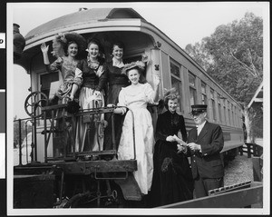 Five women standing with a railroad official on the rear of a Denver & Rio Grande Western car in turn-of-the-century costume, Knott's Berry Farm, ca.1952