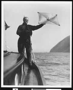 A man on a boat holding a caught fish and a kite, ca.1930