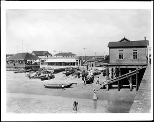 View of the Newport Beach pier, showing canoes and a man stooping in the shallows, ca.1910