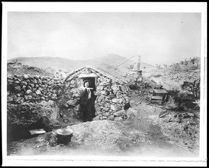 A desert miner standing in front of his stone cabin, near Randsburg, Kern County, California, ca.1900-1920