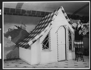 Woman in costume posing by a small gingham covered house at a Chamber of Commerce exhibit, ca.1930