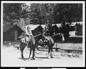 A woman and a man on horseback in front of a row of cabins, ca.1930