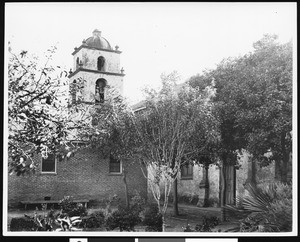 Corner of San Buenaventura Mission garden showing a side entrance, bell tower, trees and a brick wall, ca.1902