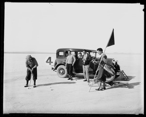 Men looking at a golfer set up a shot near an automobile