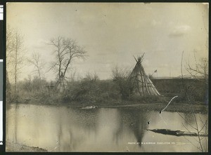 Indian camp on a river with a flag in the background, ca.1900