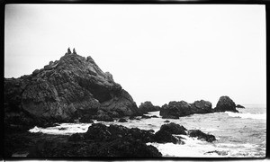 Large rocks along the ocean, showing two people at the peak of the tallest rock with one person slightly raising arm and larger landmasses