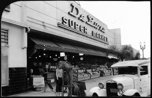 Exterior view of the DeLuxe (De Luxe?) Super Market showing produce stands and a Canal Health Food Company car, ca.1940