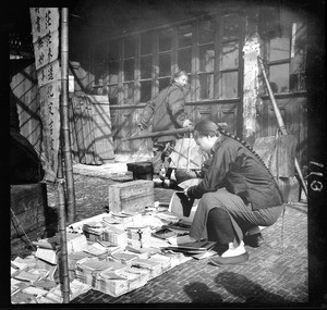 Periodical vendor setting out his wares on a cobblestone street in China, ca.1900