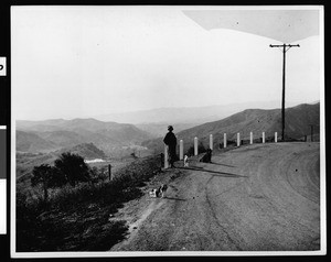 View looking west from the summit of Modjeska Grade, showing a woman and her dogs at the road-bend, El Toro, 1939