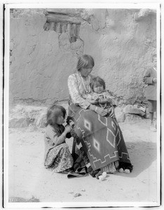 Navajo Indian mother and two children sitting in front of an adobe dwelling, ca.1900