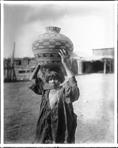 Apache Indian girl carrying an olla (a water basket) on her head, ca.1900