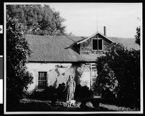 Exterior view of the Miguel Parra adobe, showing a woman with two dogs, ca.1930