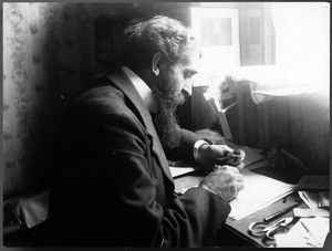 George Wharton James at his desk with a small bird, "Scraggles", perched on his finger, ca.1890-1905