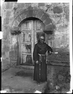 Franciscan priest standing in front of a tomb at Mission Santa Barbara, 1898