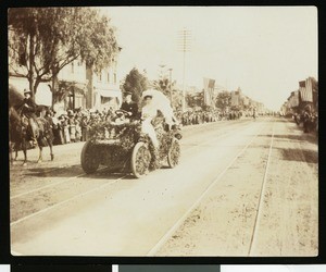 Man and woman in a foliage-adorned automobile in what appears to be a parade