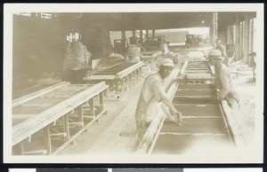 Men standing near tables in the interior of an unidentified factory, ca.1920