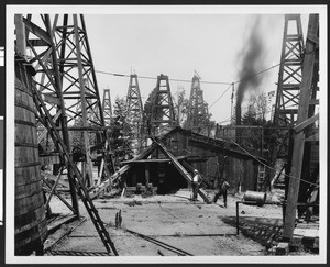 Two men standing beside a wooden triangular structure surrounded by derricks, 1904