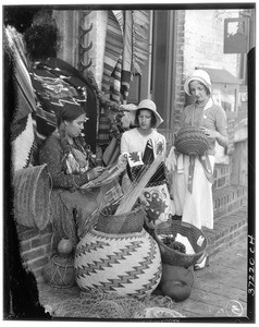 Two women standing next to a Native American woman weaving baskets