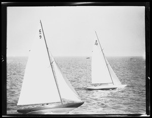 Close-up view of two sailboats on ocean with numbered sails, showing small ship in background