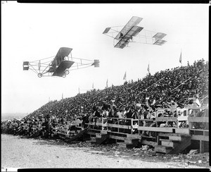 Composite photograph of two early-model biplanes passing by bleachers filled with spectators at the 1910 Dominguez Field Air Meet, January 1910