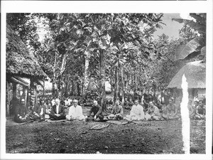 Royal Samoan council sitting on the ground amongst villagers in a Samoan village, ca.1900