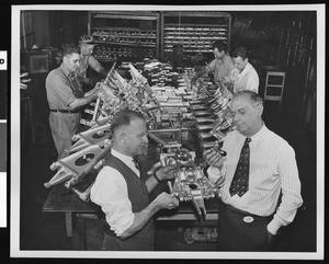 Interior view of an unidentified factory, with two suited men inspecting metal parts, while workers are on an assembly line in background, ca.1942