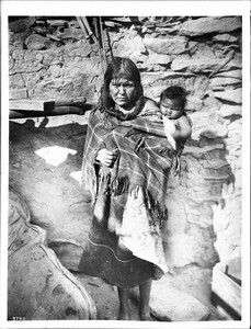 Hopi Indian mother carrying her baby on her back outside an adobe dwelling, ca.1900