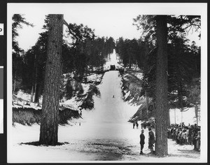 Ski jump in the mountains, showing people lining the ski slope to watch a skier, ca.1930