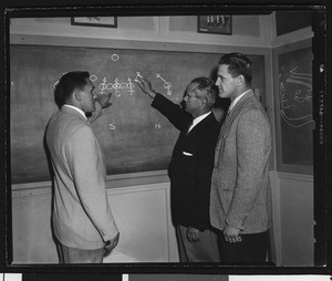 University of Southern California football coaches pointing at a chalkboard with football plays diagrammed on it, USC campus, 1951