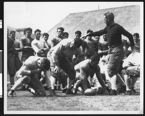 University of Southern California football coach Howard Jones in a coaching situation, pointing, players in formation. USC player John Wayne is in the background