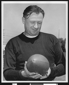 University of Southern California head football coach Jeff Cravath, shot to waist, dark sweatshirt, holding a football and looking at it. Bovard Field. 1949