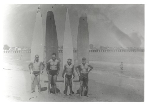 Unidentified, Alex Hokamp, Harry Mayo, Fred Hunt standing in front of surfboards on Cowell Beach