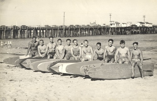 Club surfers sitting in front of surfboards: Don "Mighty Bosco" Patterson, Rich Thompson, Alex Hokamp, Blake Turner, Bill Grace, Dave "Buster" Steward, Fred Hunt, Harry Mayo, Alex (Pinky) Pedemonte, Tommy Roussel, Harry Murray at Cowell Beach