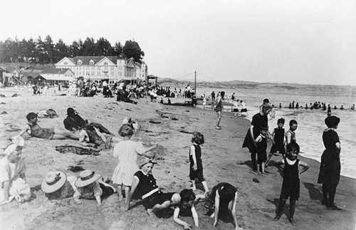 Sunbathers, swimmers, and children playing at the Capitola City Beach, with Hotel Capitola