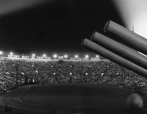 Large stadium celebration in support of the war effort. Large letters are illuminated at the far top edge of the stadium: "VICTORY MUST BE PAID FOR. BUY VICTORY BONDS." Three canons protrude from the right side and the stadium is packed with supporters holding lights