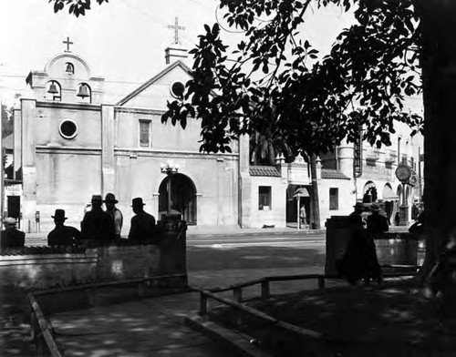 The Front of the Plaza Church (Main Street side showing Plaza shoe shine on south side of church and La Azteca on north side). Also pictured is part of the Plaza with men sitting on benches facing the Church