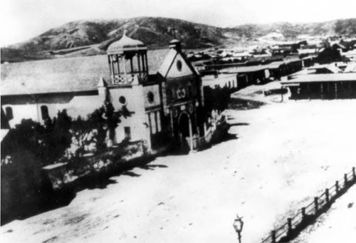 View of church showing south side and front facade with gothic style belfry, portion of fence around the plaza in lower right hand corner. Sonoratown in background with hills